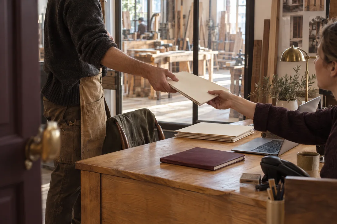Deux collègues se transmettent un dossier dans le bureau d’un atelier artisanal.